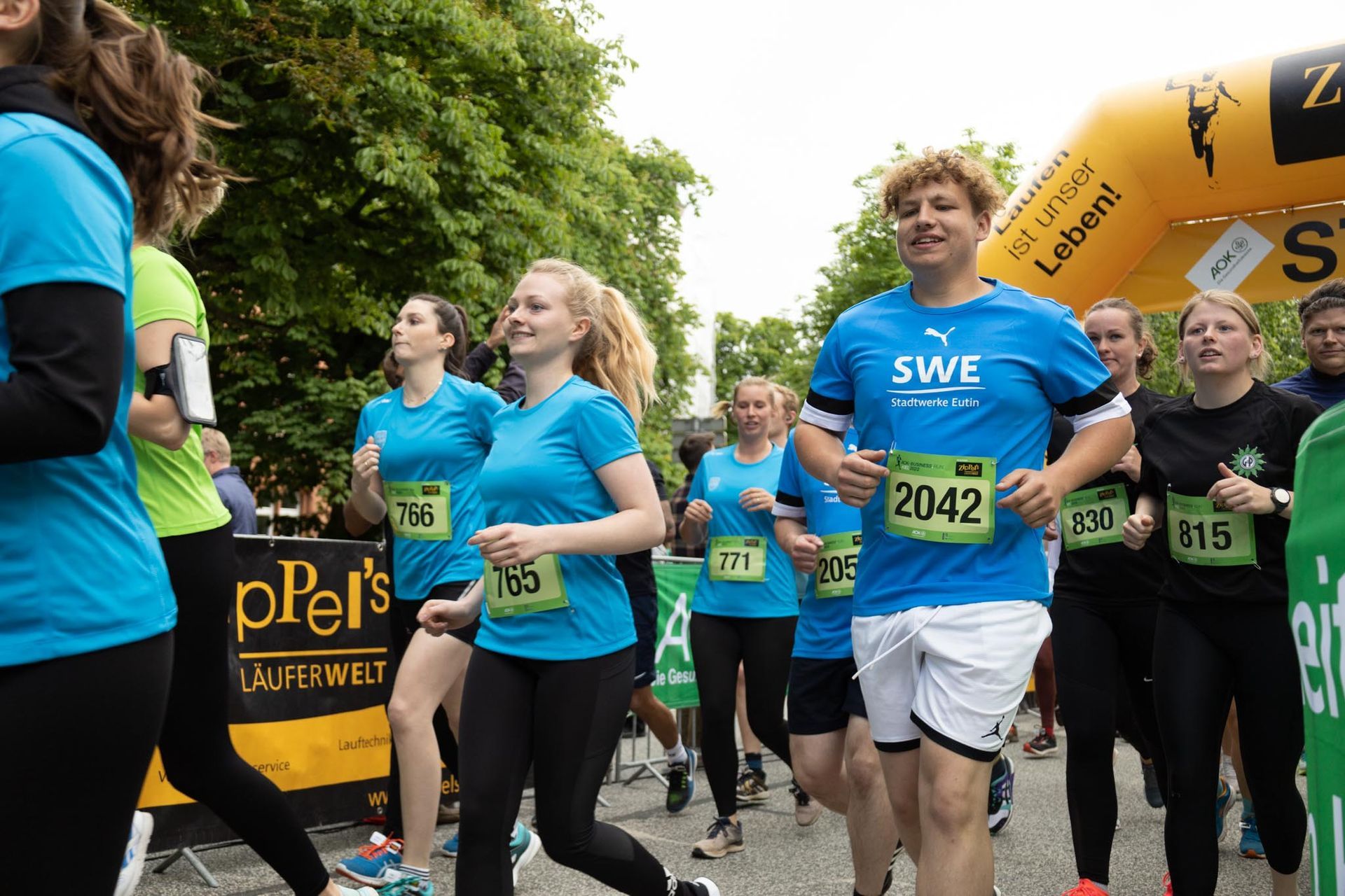 Läufer bei einem Wettlauf, einige mit Startnummern, unter einem gelben Bogen.