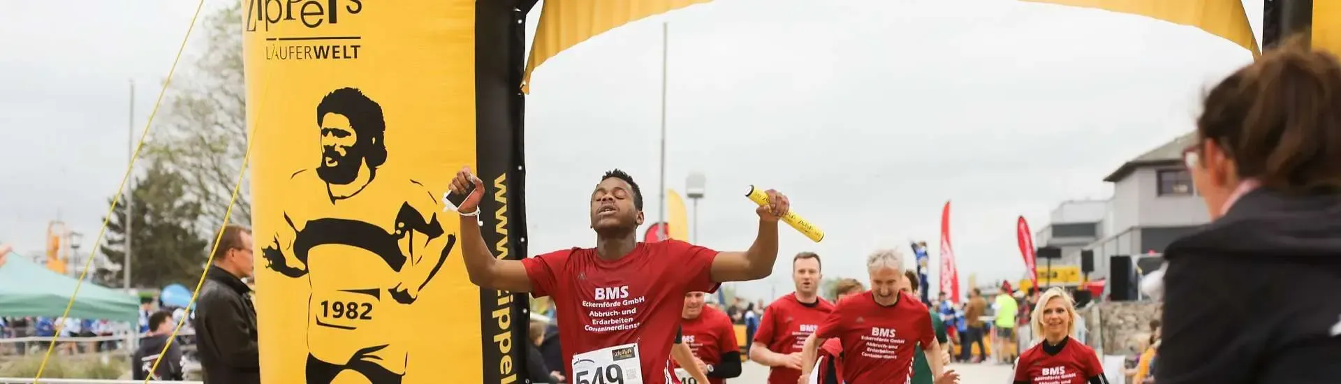 Ein Läufer in einem roten Hemd reißt nach dem Überqueren der Ziellinie die Arme zum Siegesjubel hoch; im Hintergrund ist ein gelbes Banner zu sehen.