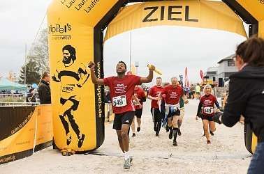 Ein Läufer überquert die Ziellinie unter einem mit „ZIEL“ beschrifteten Torbogen. Andere folgen ihm, Zuschauer stehen an den Seiten.