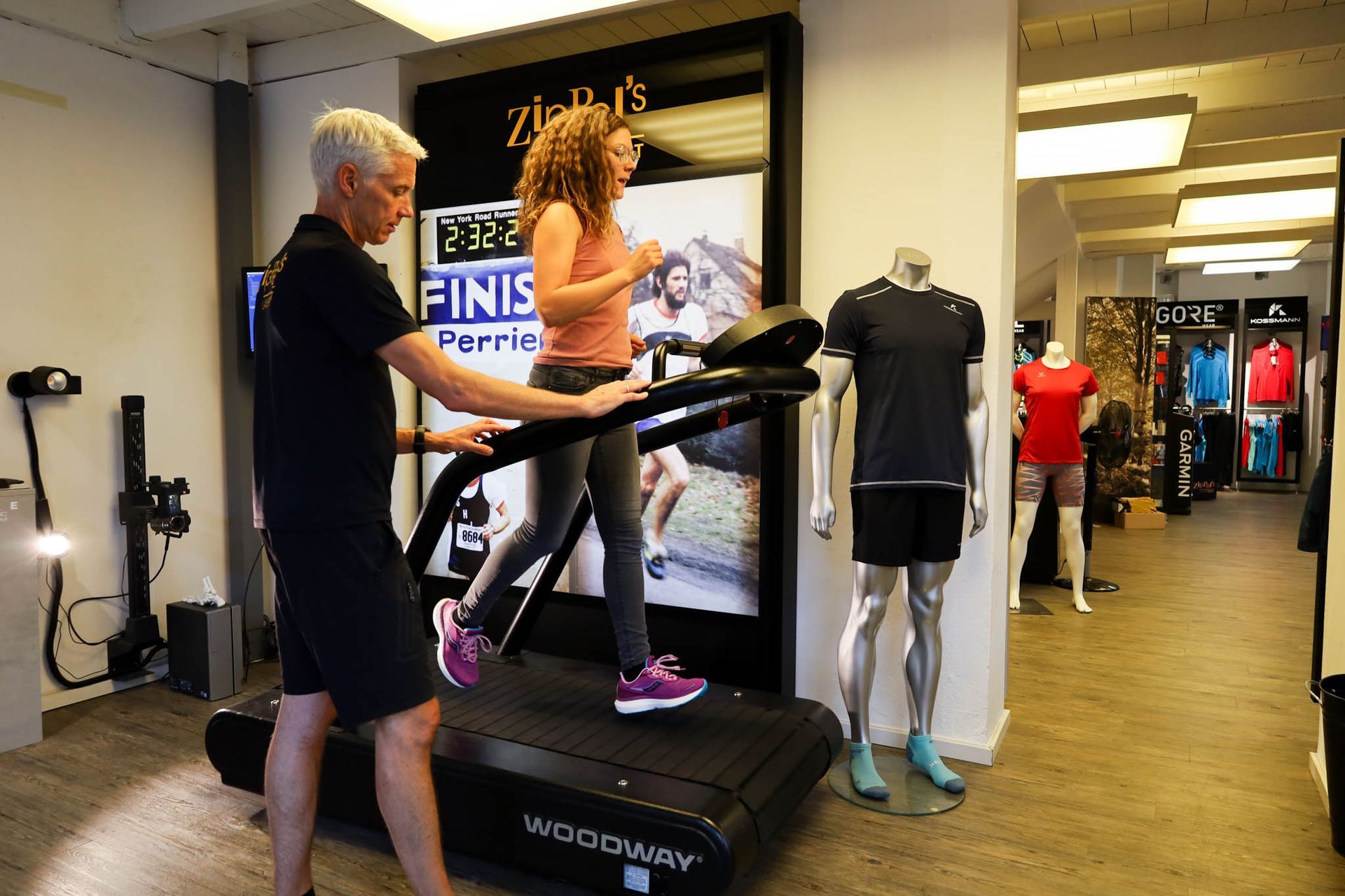 Eine Frau läuft auf einem Laufband und wird dabei von einer Person in einem Geschäft für Sportbekleidung unterstützt.