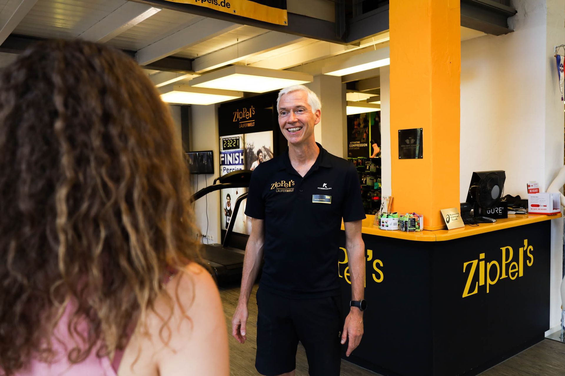 Ein Mann in schwarzem Hemd lächelt eine Frau in einem Fitnessstudio an. Das Logo von Zip'r prangt auf der Theke, eine orangefarbene Akzentwand setzt einen schönen Akzent.