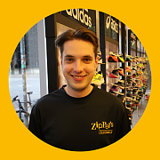 Ein Mann in einem schwarzen Zip-T-Shirt lächelt in einem Schuhgeschäft; die Logos von Adidas und Asics sind sichtbar.