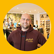 Ein lächelnder Mann in einem weinroten Kapuzenpullover befindet sich in einem Laufgeschäft. Im Hintergrund sind Schuhe und Sportbekleidung zu sehen.