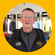 Mann mit Brille und schwarzer Jacke lächelt in einem Sportgeschäft, gelber Hintergrund.