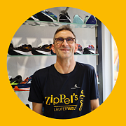 Ein lächelnder Mann in einem Schuhgeschäft trägt ein schwarzes Hemd mit der Aufschrift „Zippel's LAUFERWELT“. Hinter ihm sind Schuhe ausgestellt.