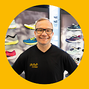 Ein Mann mit Brille lächelt und steht in einem Schuhgeschäft, in dem Laufschuhe ausgestellt sind.