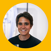 Lächelnde Person mit kurzen dunklen Haaren und schwarzem Hemd in einem Raum mit gelbem Kreishintergrund.