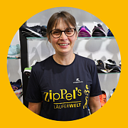 Eine lächelnde Frau mit Brille und einem T-Shirt mit dem Logo „zipPel's LÄUFERWELT“ in einem Schuhgeschäft.