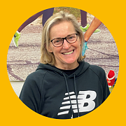 Frau mit Brille lächelt und trägt einen New Balance Hoodie; im Hintergrund sind Laufschuhe zu sehen.