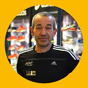 Mann in Adidas-Shirt lächelt in einem Schuhgeschäft; im Hintergrund sind Schuhauslagen zu sehen.