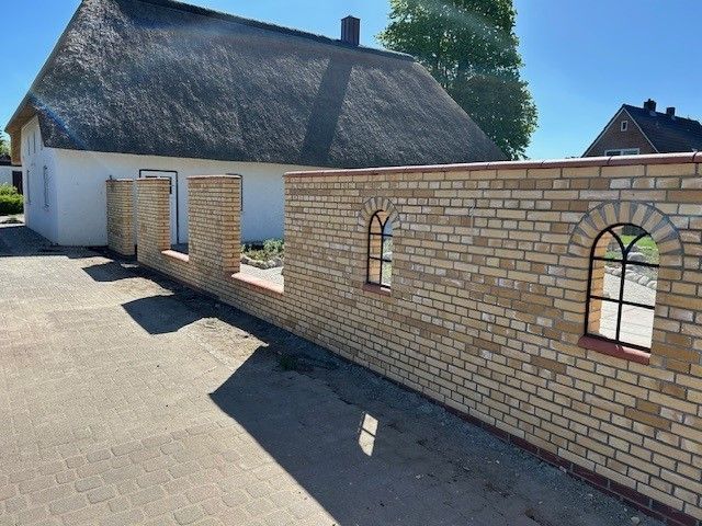 Hinter einer Backsteinmauer steht ein weißes Haus mit Strohdach