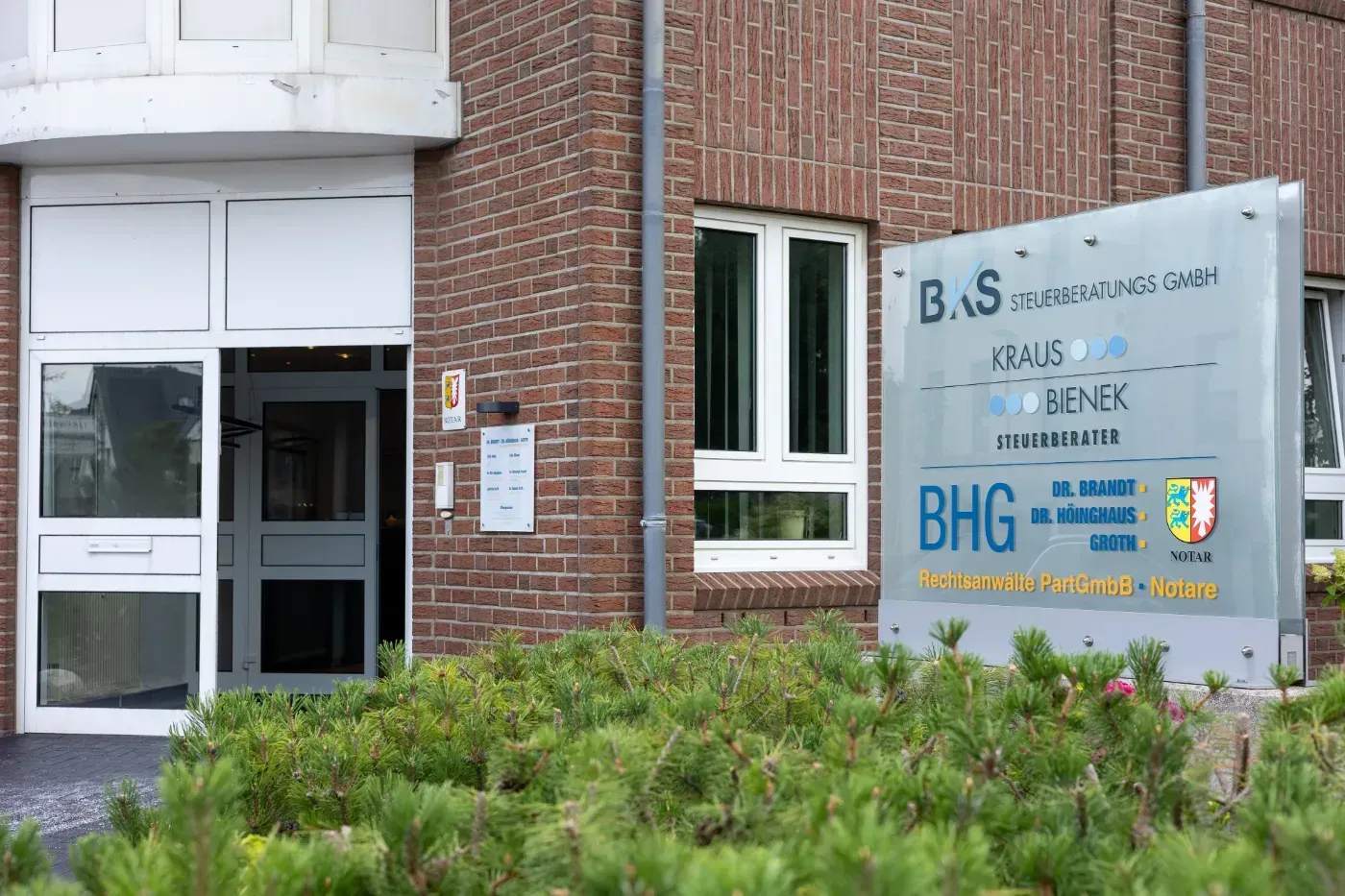 Gebäudeeingang mit einem Schild mit der Aufschrift BS und BHG neben einer Backsteinmauer und Sträuchern.