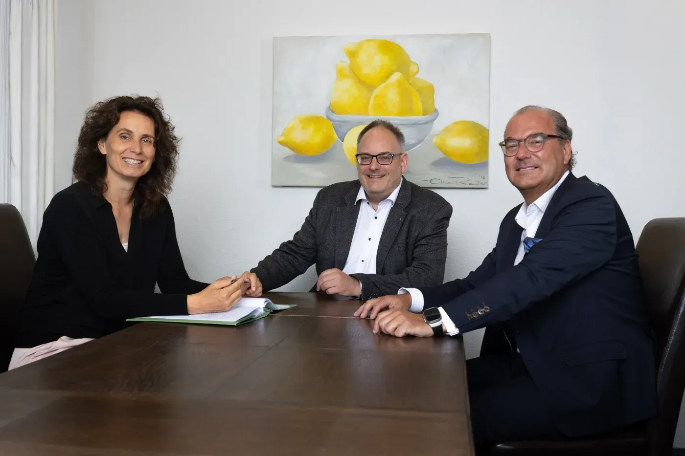 Drei Personen in Businesskleidung sitzen an einem Konferenztisch, an der Wand hängt ein Bild mit Zitronenmotiven.