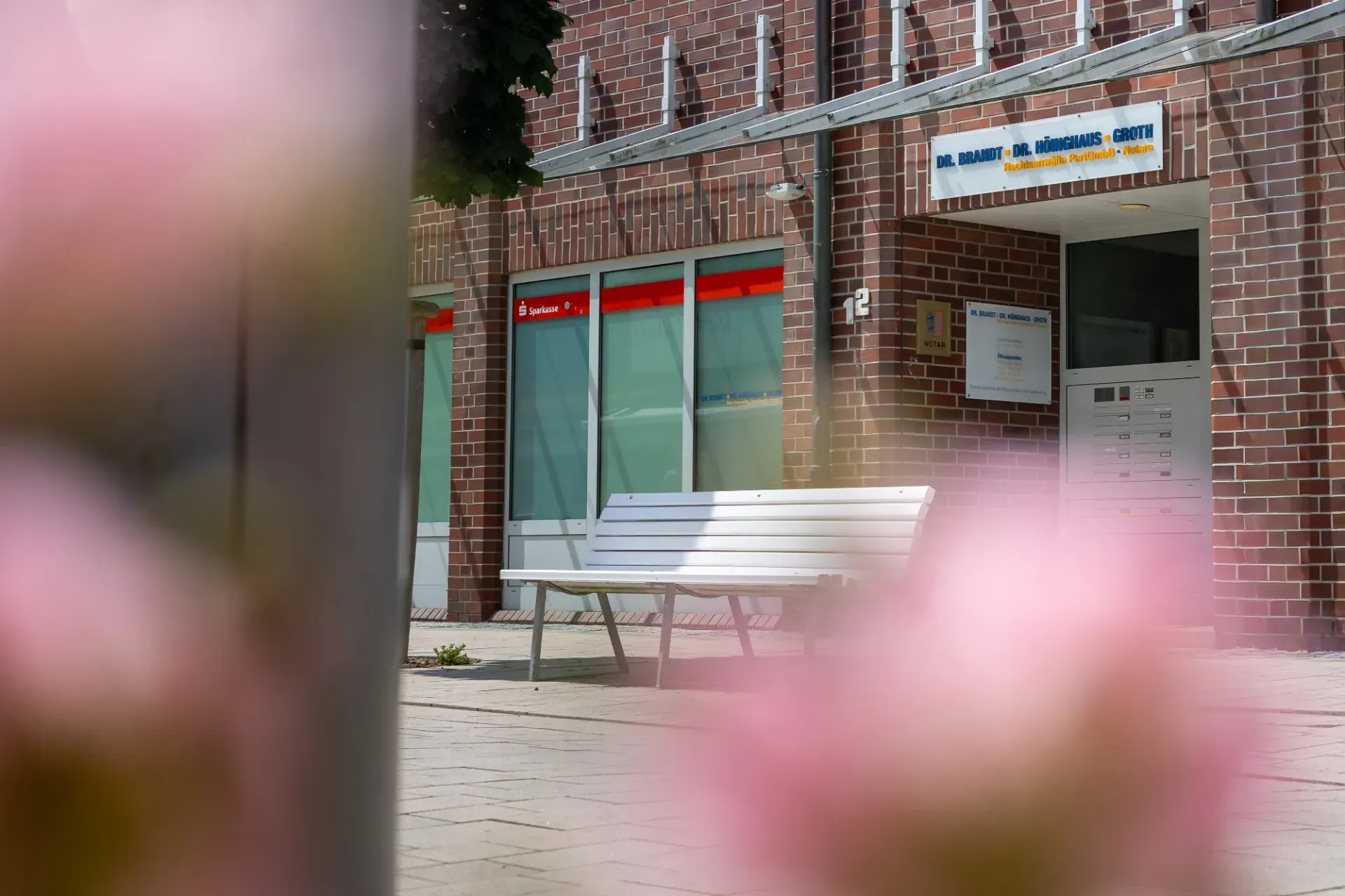 Eingang zu einem Backsteingebäude mit einer weißen Bank und im Vordergrund verschwommenen rosa Blumen