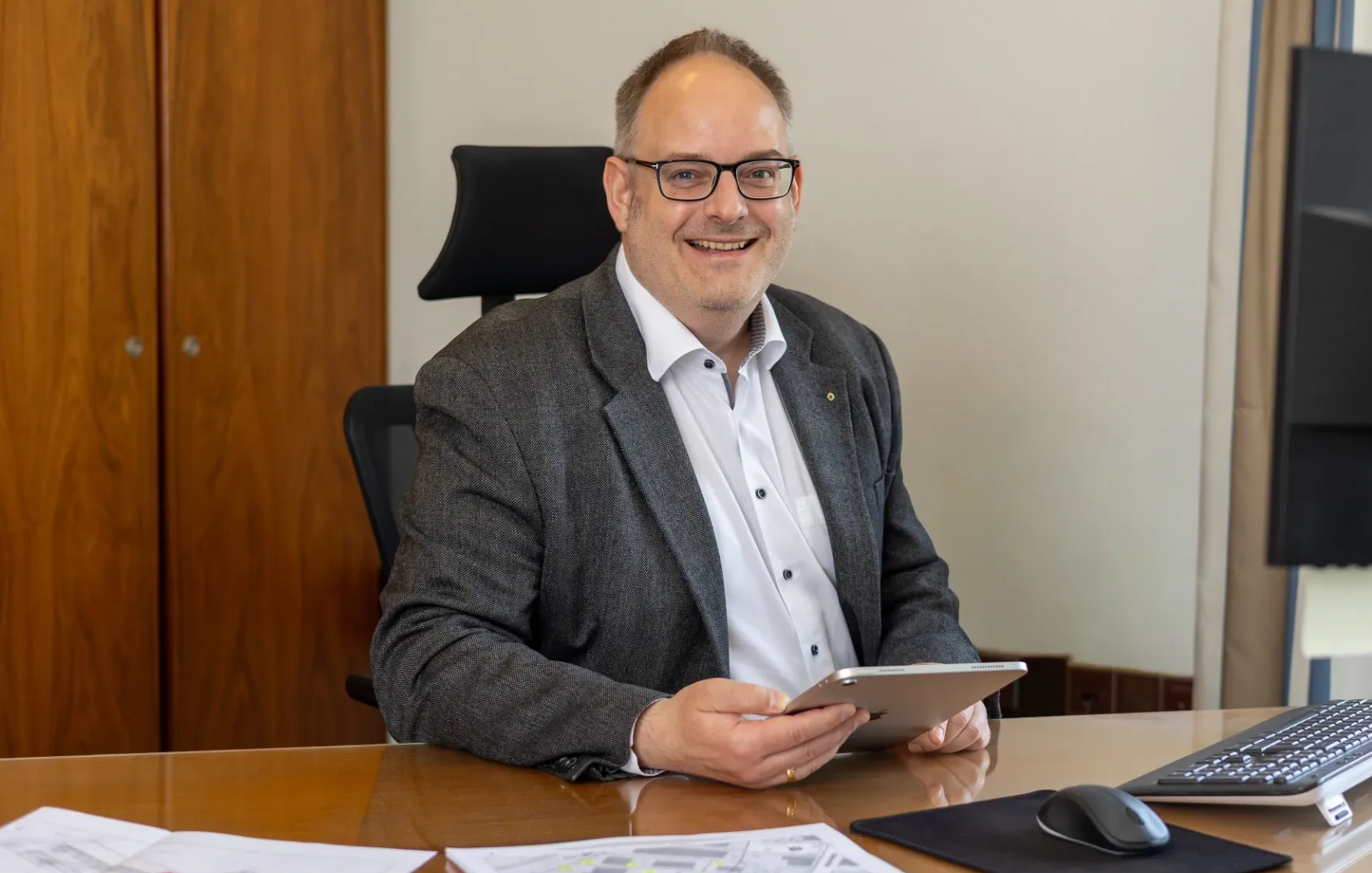 Ein lächelnder Mann in einem grauen Blazer sitzt an einem Schreibtisch in einem Büro und hält ein Tablet in der Hand.