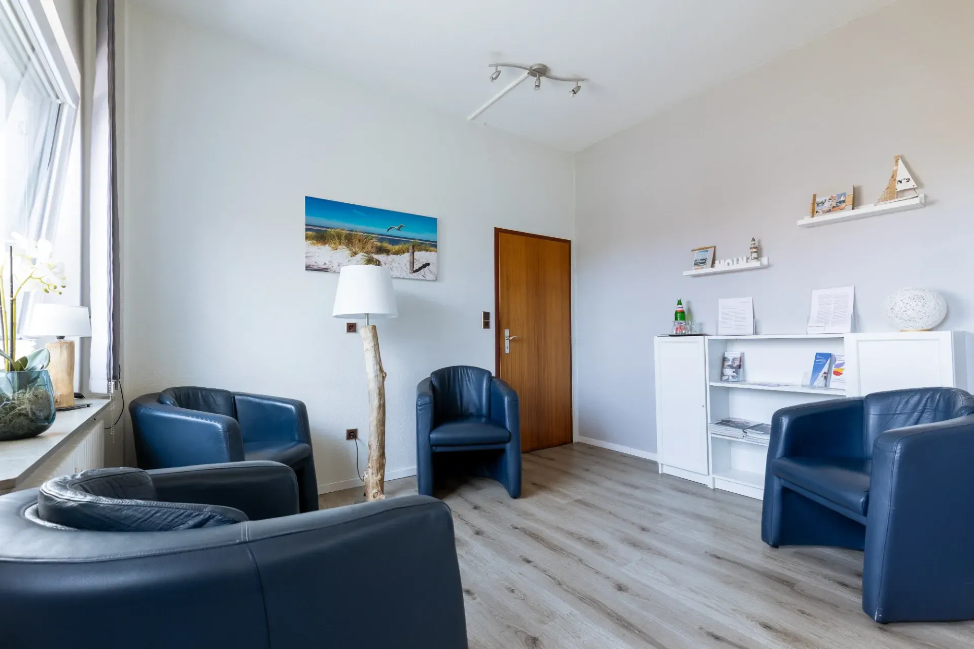 Helles Wohnzimmer mit blauen Stühlen, weißen Regalen, einer Lampe und einer Holztür auf hellem Holzboden