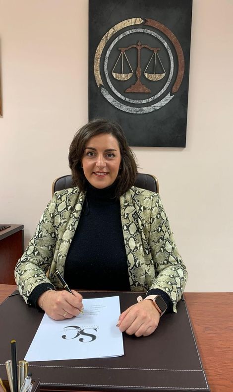 Una mujer sentada en un escritorio frente a un emblema de la justicia sostiene un bolígrafo sobre unos papeles.