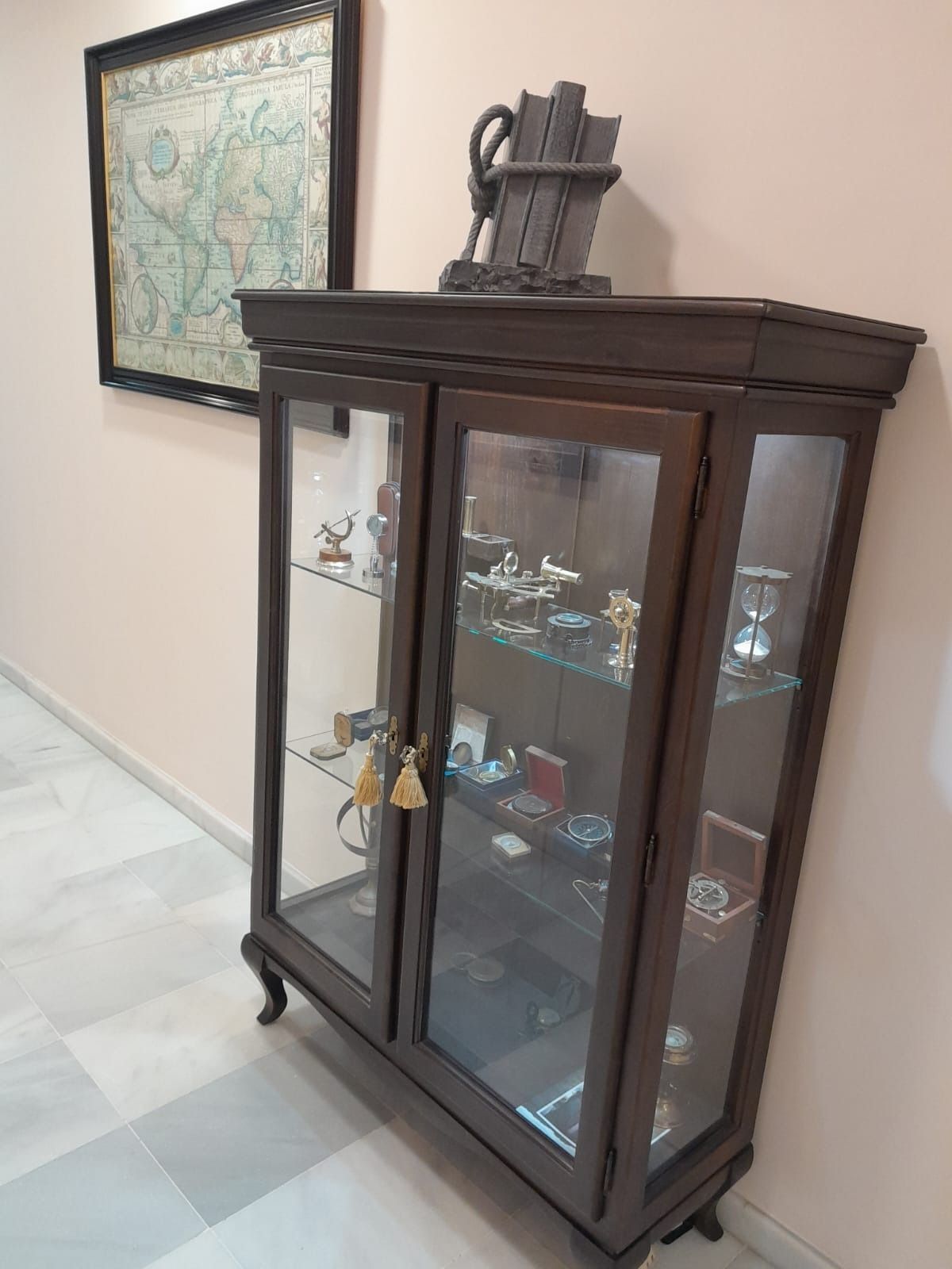 Vitrina de madera oscura con puertas de cristal que contiene objetos pequeños, junto a un mapa mural enmarcado en un pasillo.