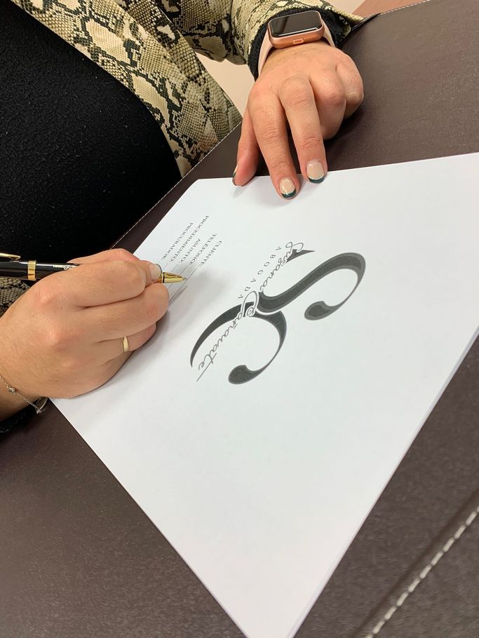 Manos escribiendo en un certificado blanco con un gran símbolo decorativo negro sobre una mesa.
