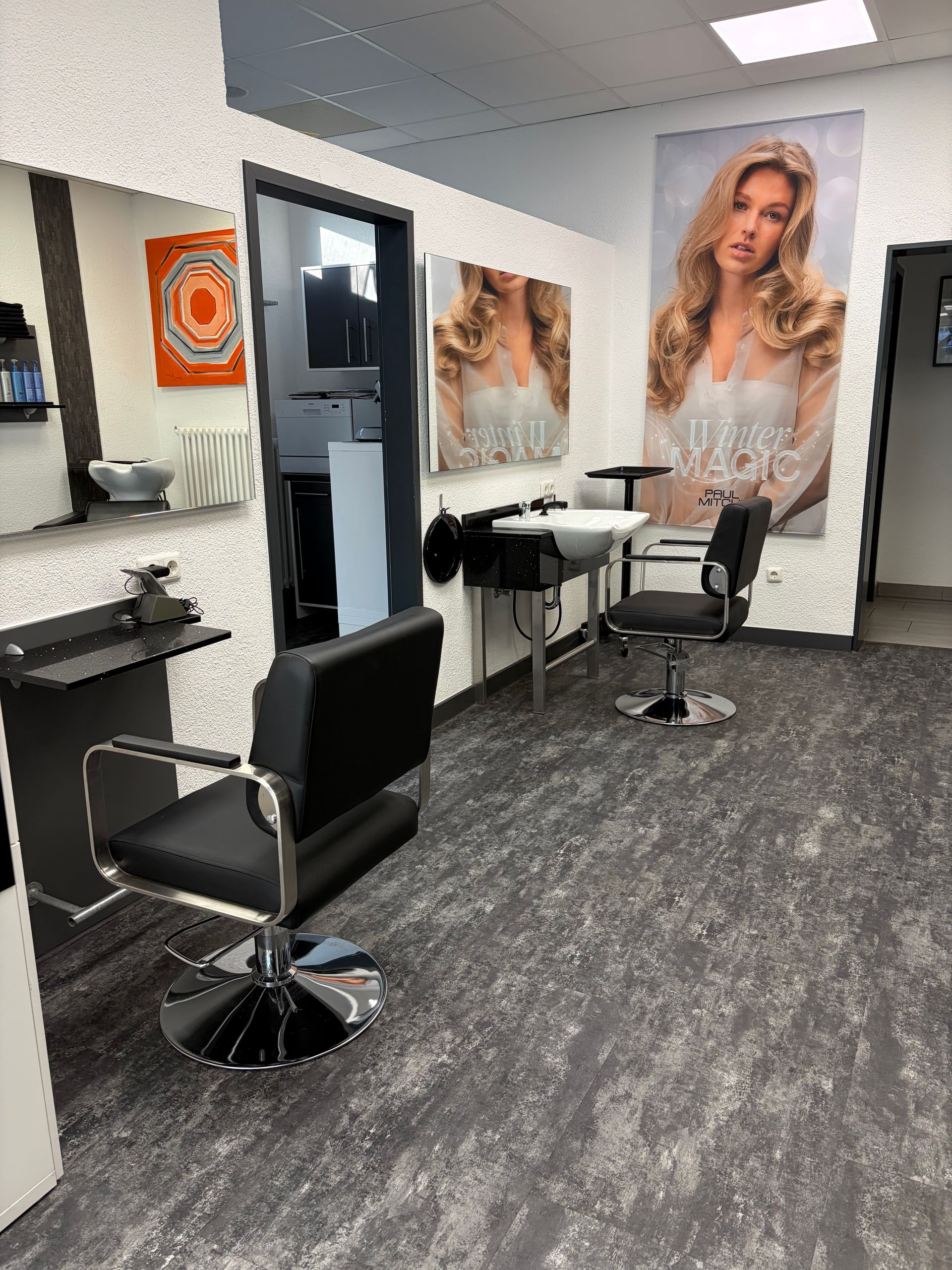Das Interieur eines Friseursalons mit zwei schwarzen Friseurstühlen vor Spiegeln und einem großen dekorativen Wandposter.