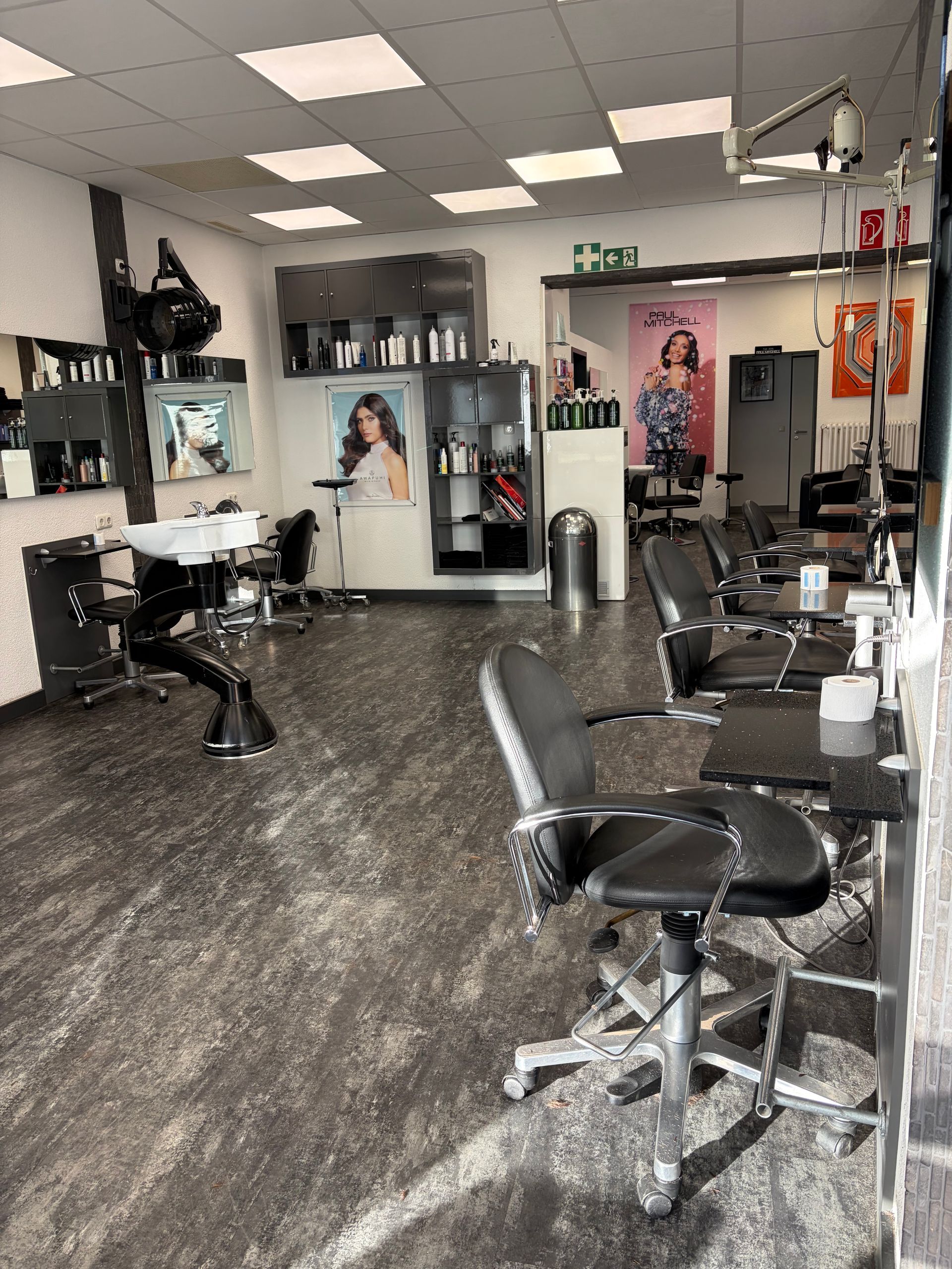Ein moderner Friseursalon mit schwarzen Stühlen, einem Waschbecken, Regalen für Produkte und heller Deckenbeleuchtung.