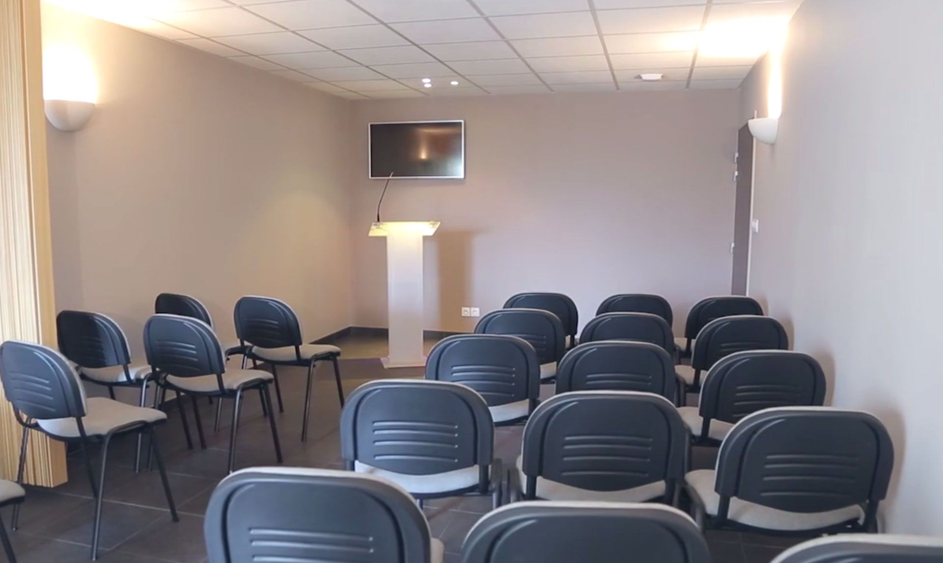 Salle de conférence vide avec des rangées de chaises bleues, un podium et une télévision fixée au mur.