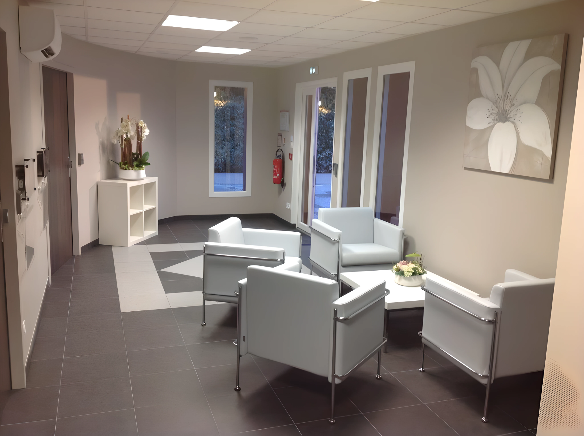 Salle d'attente moderne avec chaises gris clair, petites tables, décorations murales, fenêtres et sol carrelé