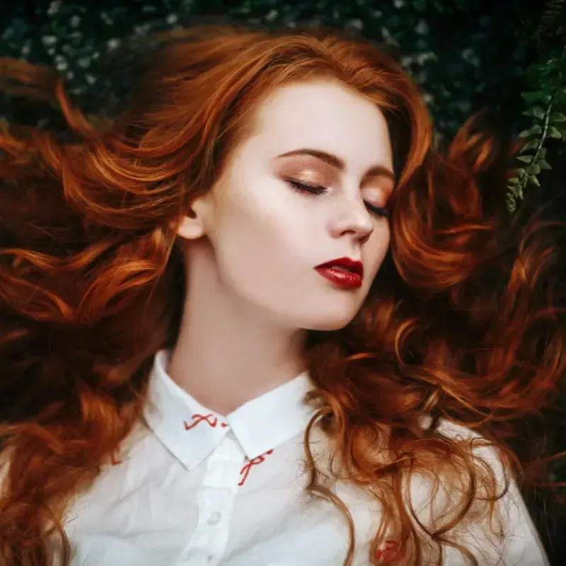 Una mujer con cabello largo y rojo yace en el follaje, con los ojos cerrados y vistiendo una camisa blanca.