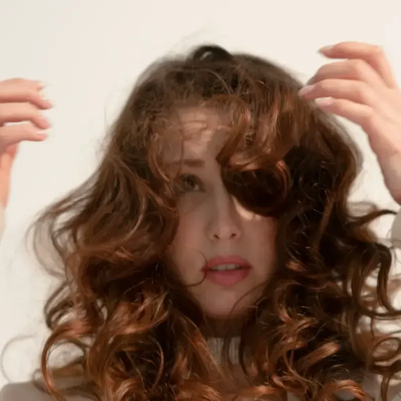 Mujer con cabello oscuro y ondulado, con lápiz labial, mirando a la cámara. Fondo gris.