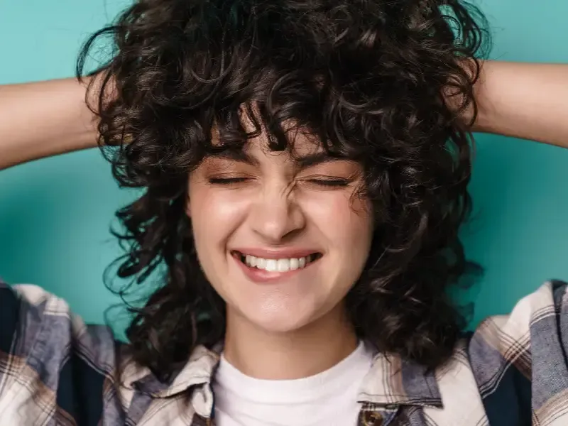Mujer con cabello rizado sonriendo, con los ojos cerrados y las manos en el cabello, sobre un fondo verde azulado.