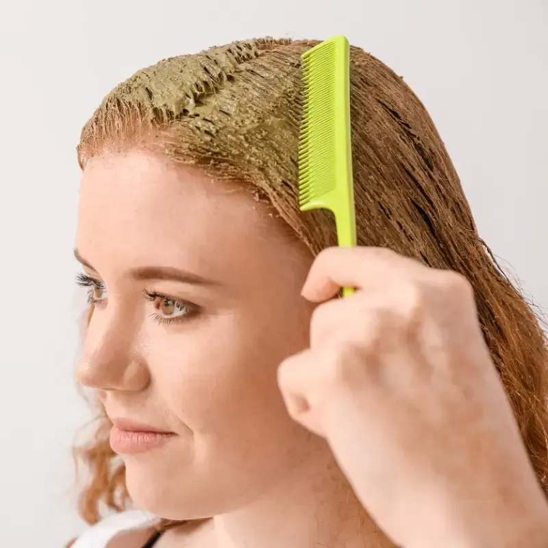 Recipiente para tinte para el cabello con aplicador, tinte en polvo y cabello naranja vibrante sobre superficie gris.