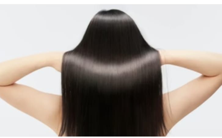 Mujer con cabello negro largo y brillante, con los brazos levantados detrás de la cabeza, sobre un fondo blanco.