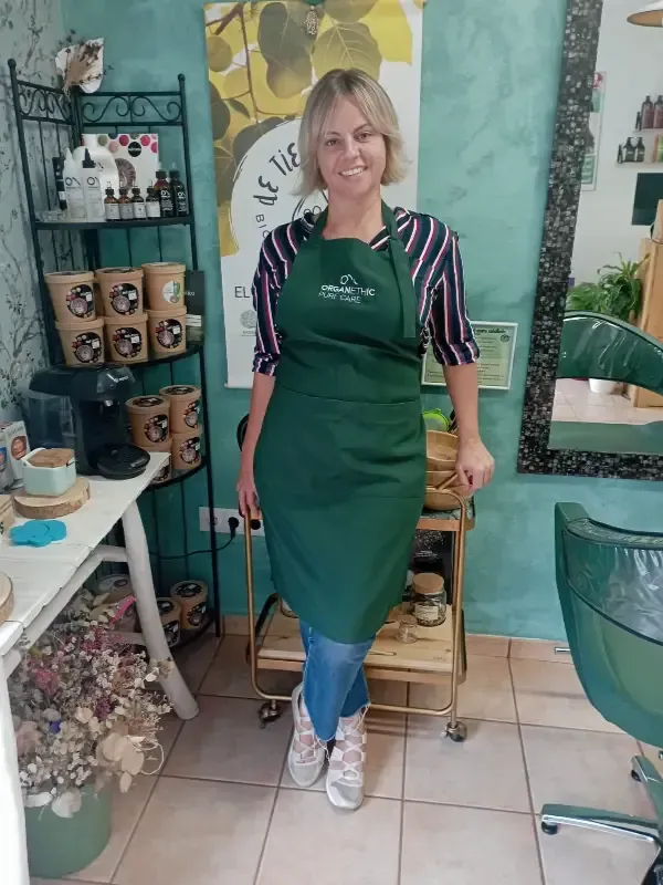 Cristina Ramírez con delantal verde y camisa de rayas, sonriendo en su salón con productos y decoración.
