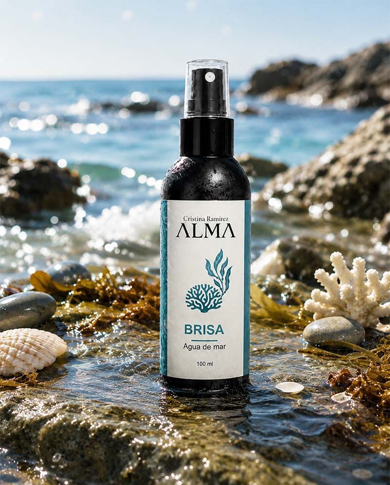Botella de spray ALMA Brisa en una playa rocosa con conchas marinas y olas del océano al fondo.