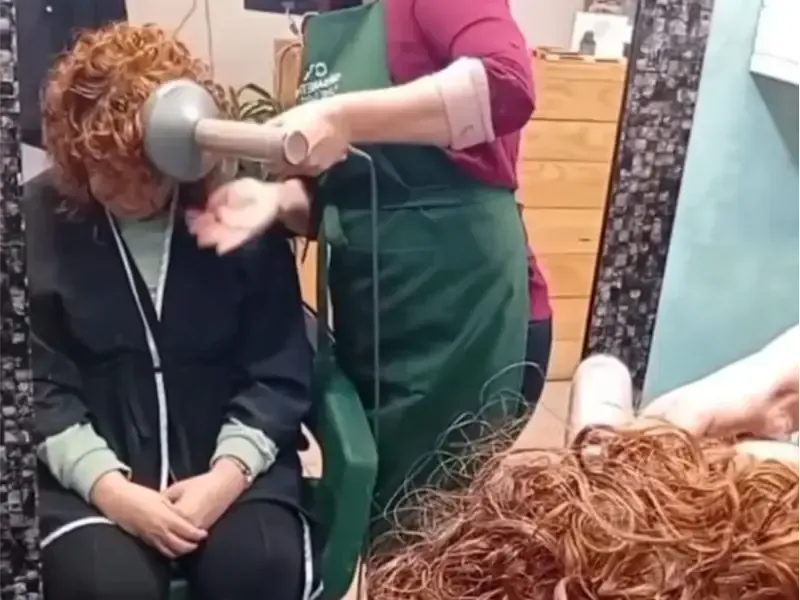 A una mujer sentada en una silla de salón le secan el cabello con un secador de mano alguien con un delantal verde.