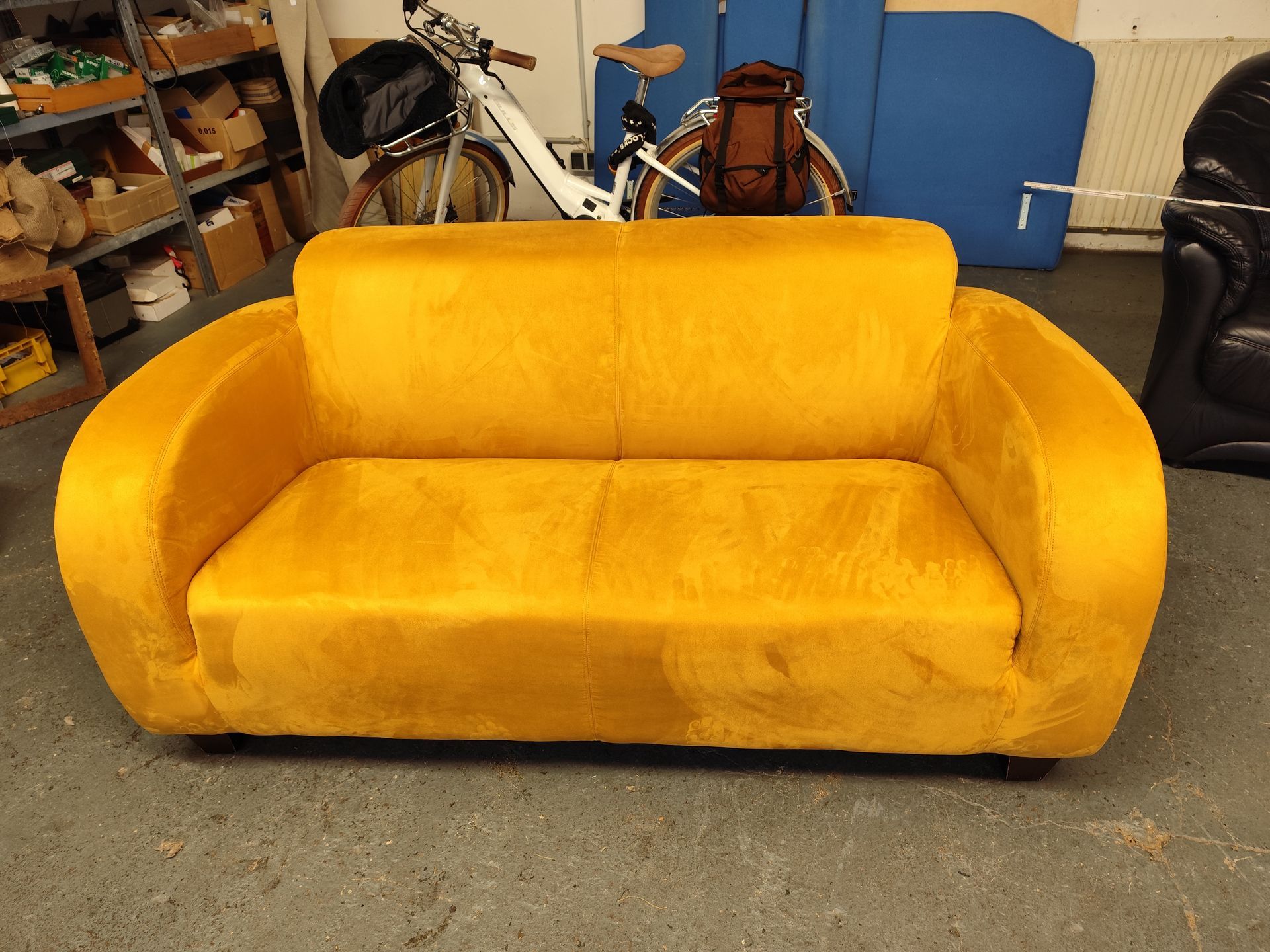 Senfgelbes Zweisitzer-Sofa. Im Hintergrund sind ein Fahrrad und Möbel in einer Werkstattumgebung zu sehen.
