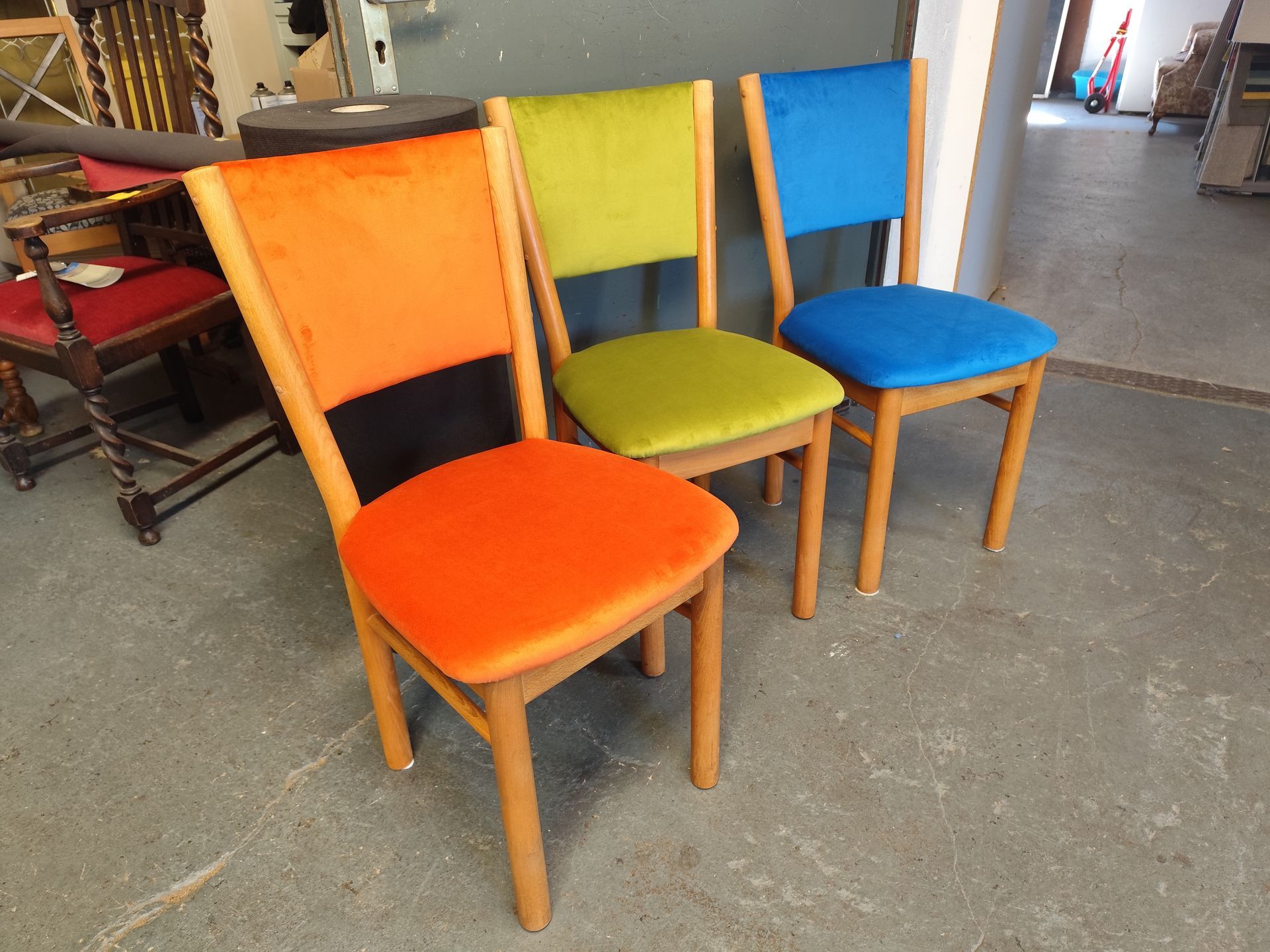 Drei Holzstühle mit orangefarbenen, grünen und blauen Sitz- und Rückenlehnen stehen auf einem Betonboden.