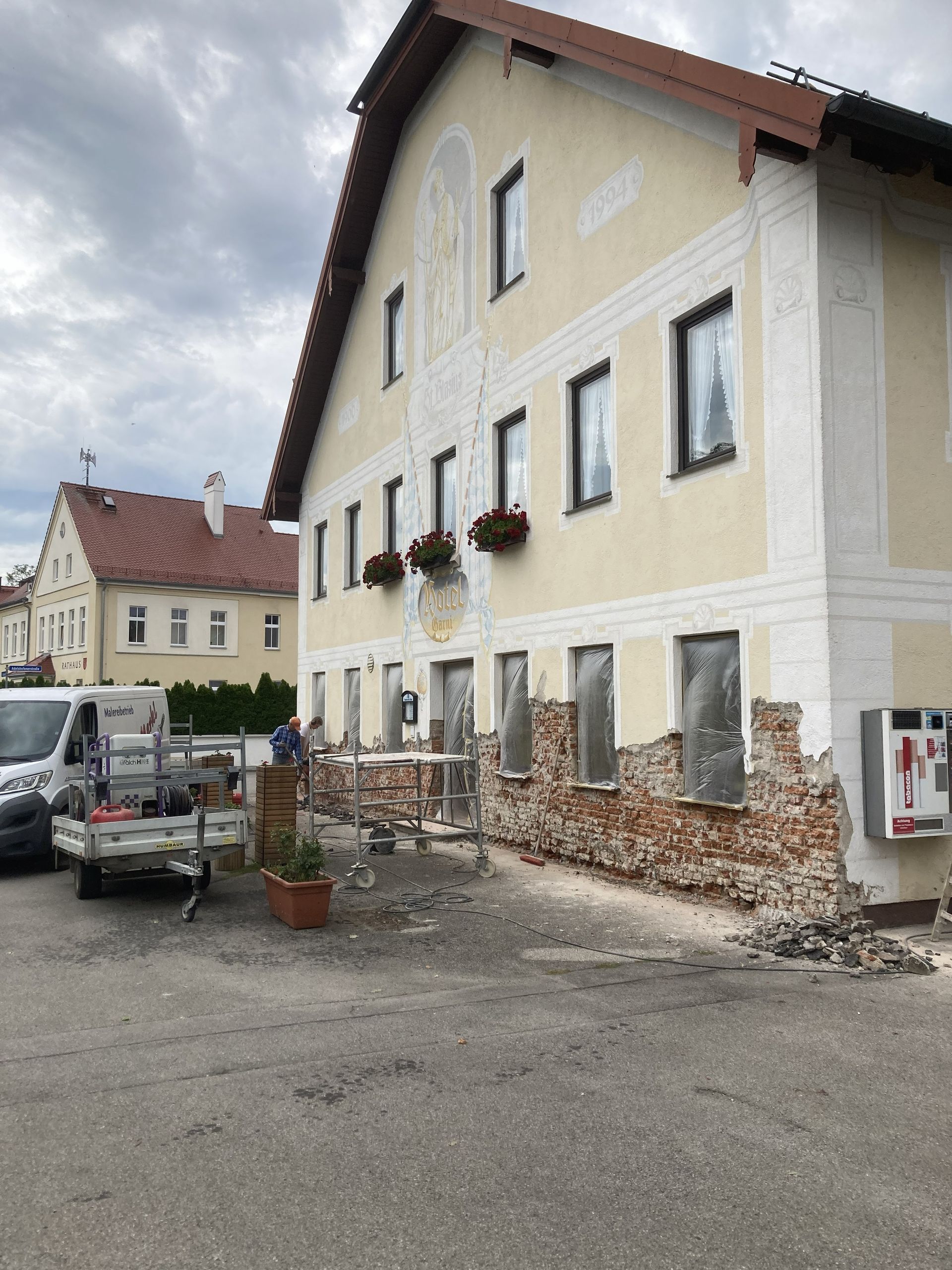 Ein Maler auf einer Leiter trägt graue Farbe auf eine Wand. Er trägt einen blauen Overall und eine Mütze. Weiße Eimer und eine Farbrolle sind zu sehen.