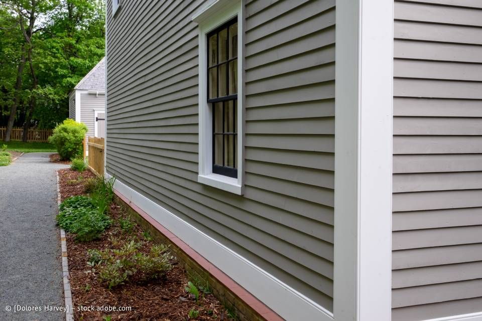 Graue Schindelverkleidung an einem Haus mit einem weiß getönten Fenster und einem Blumenbeet entlang eines Kieswegs.