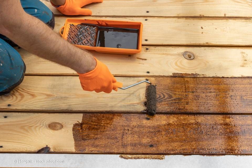 Person mit orangefarbenen Handschuhen beizt Holzbretter mit einer Rolle und verwendet dabei eine Schale mit brauner Beize.