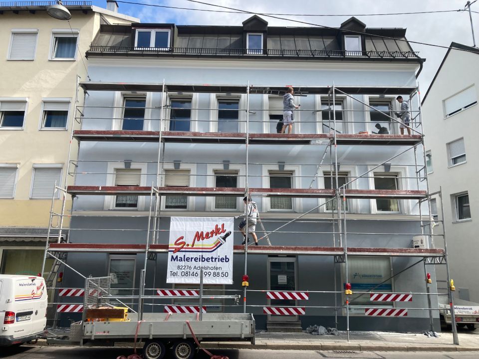 Gebäudeaußenseite wird mit Gerüst renoviert; Arbeiter auf den Ebenen streichen hellblaue Fassade.