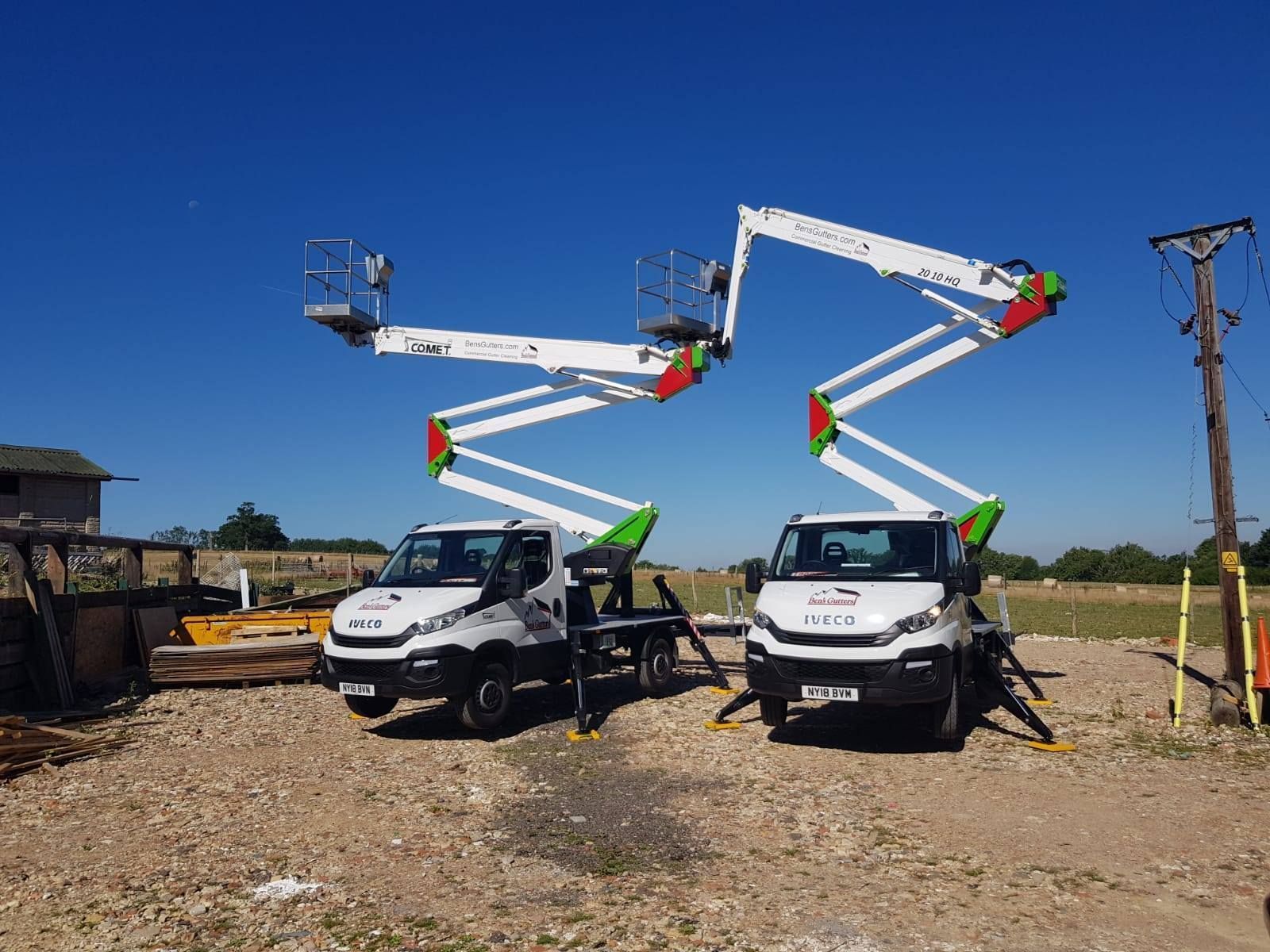 Two white utility trucks with raised bucket lifts in a gravel lot under a clear blue sky