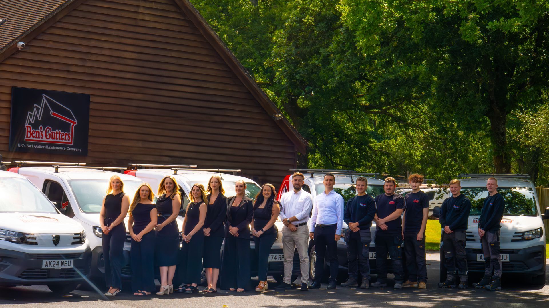 Group of people standing outside vans and a wooden building in a sunny wooded area