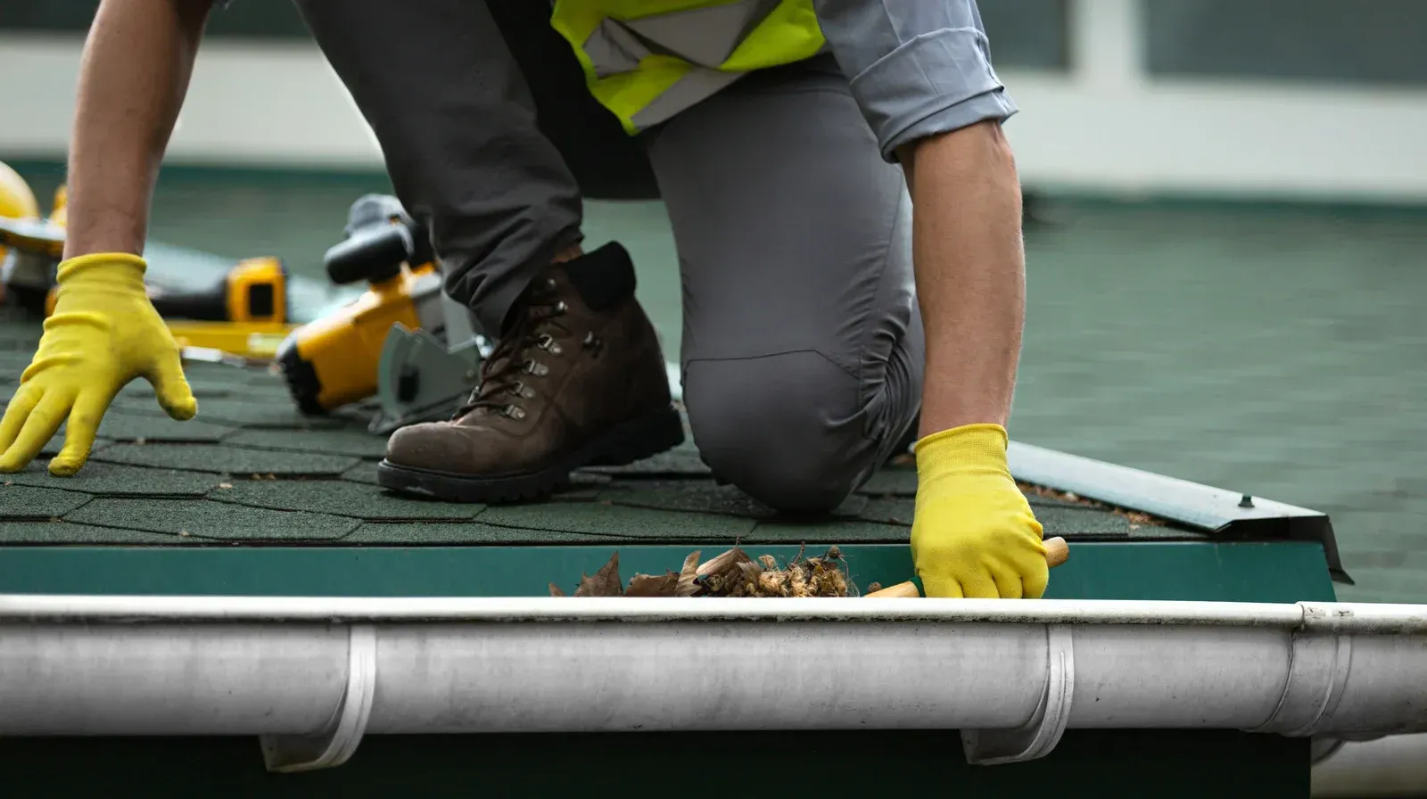 Worker in yellow gloves kneeling on a roof, cleaning debris near a gutter with tools nearby