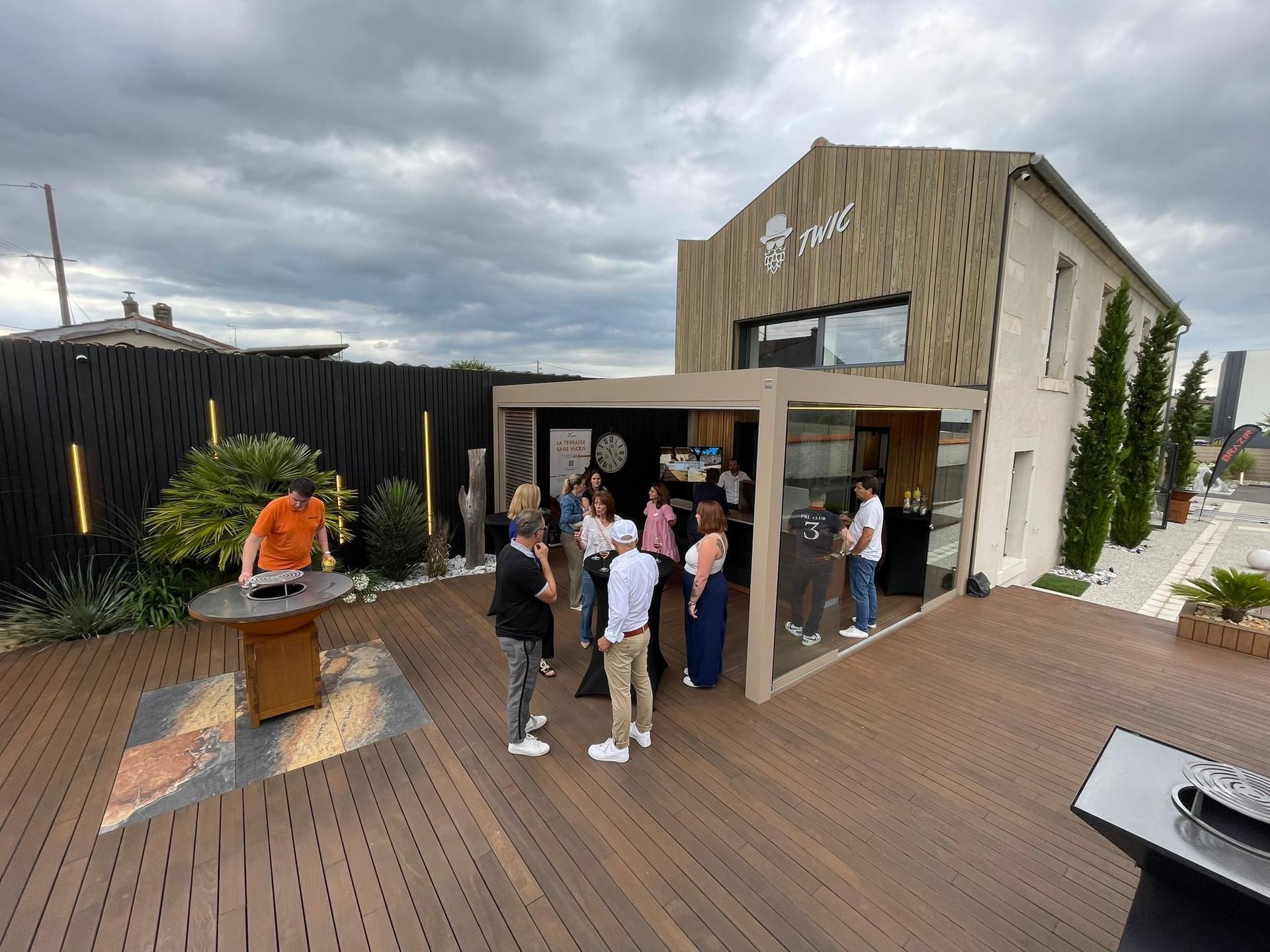 Des gens se rassemblent devant un bâtiment moderne doté d'une terrasse en bois ; un homme fait des grillades.
