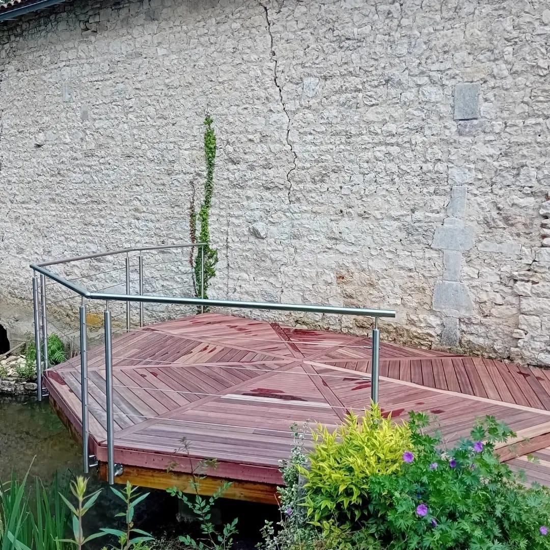 Terrasse en bois avec rambarde métallique au bord de l'eau, mur de pierre en arrière-plan.