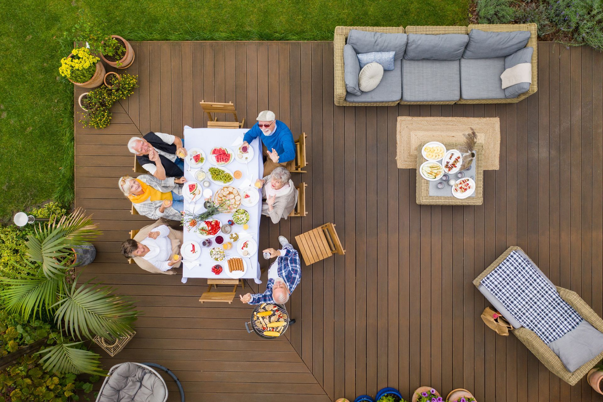 Vue aérienne de personnes assises autour d'une table sur une terrasse, profitant d'un repas, avec du mobilier de jardin et des plantes à proximité.