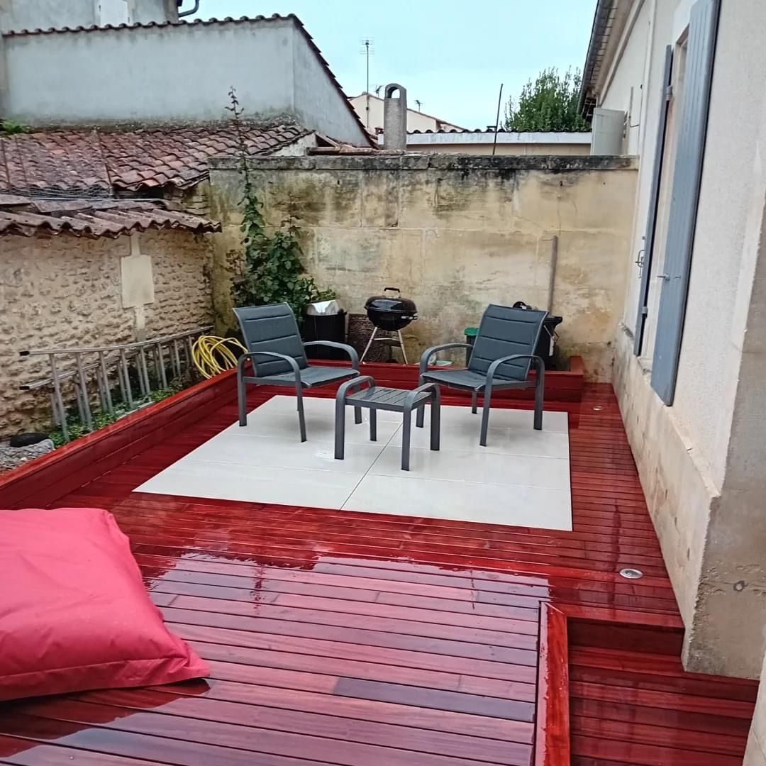 Terrasse en bois teinté en rouge avec mobilier de jardin, comprenant des chaises et un repose-pieds, et un petit barbecue, dans une cour.