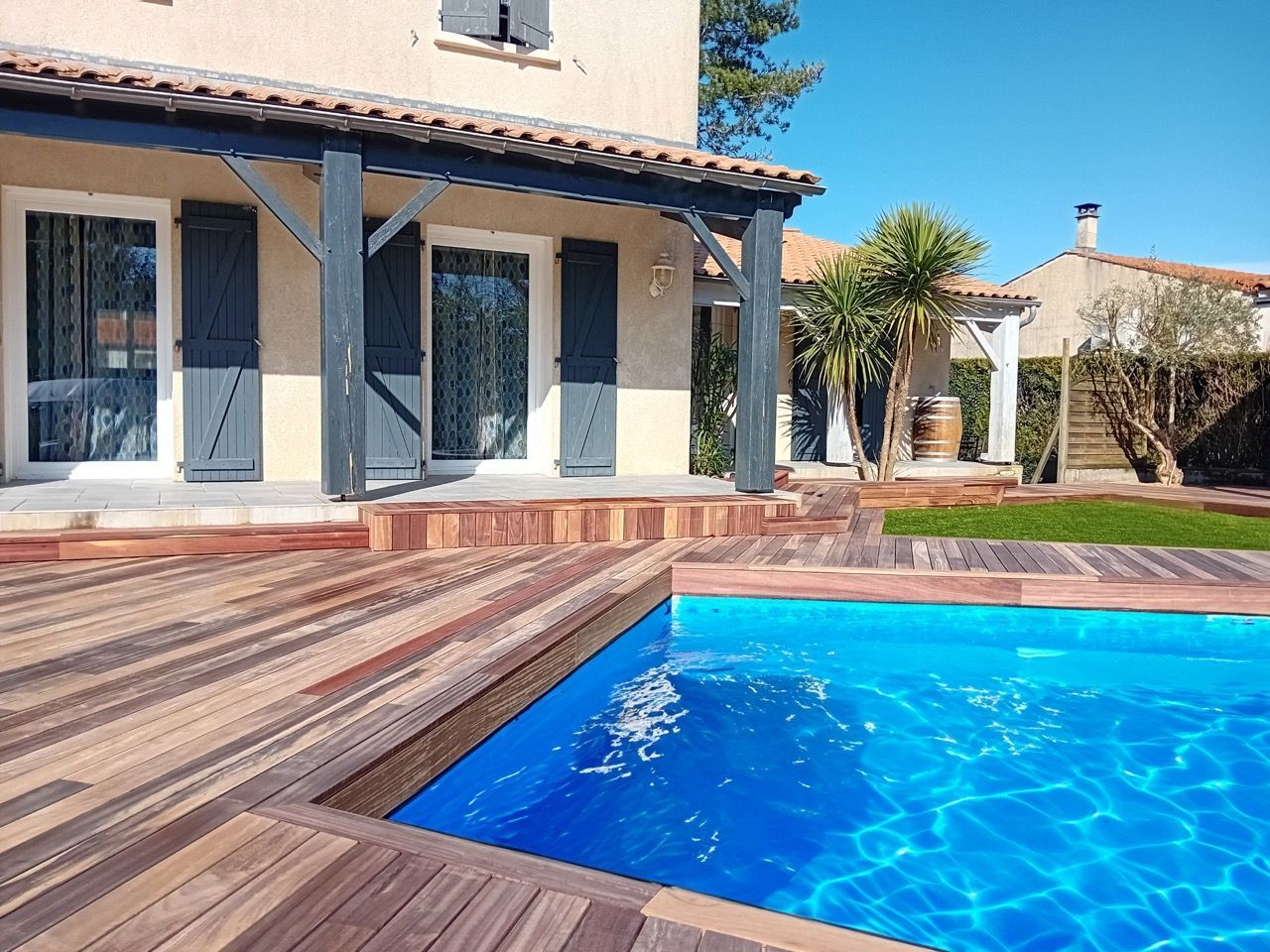 Piscine jouxtant une terrasse en bois et une maison aux volets bleus.