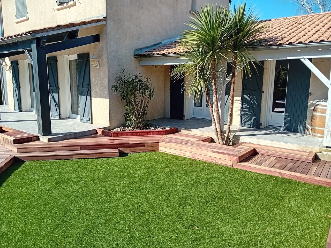 Vue extérieure d'une maison avec une pelouse verte, une terrasse en bois et un palmier.